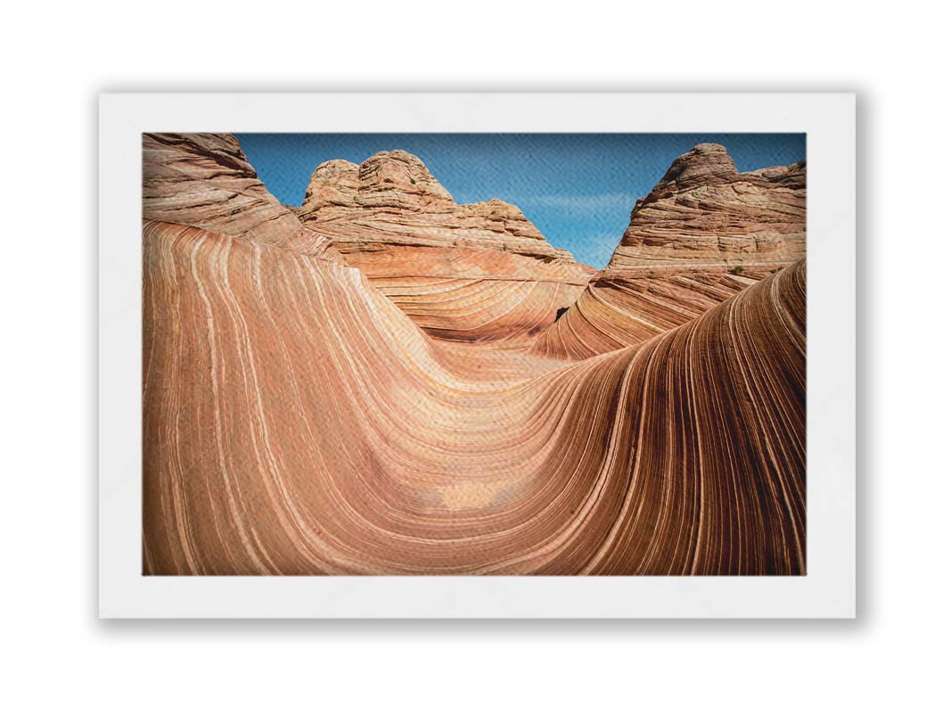 A photo of the "Wave" rock formation at Coyote Buttes North in Utah. The wavy sandstone layers create rolling waves where they have been carved out of the mountainside. The orange hues of the stone are sharply contrasted by the vibrant blue of the sky. Printed on canvas and framed.