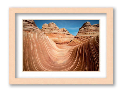 A photo of the "Wave" rock formation at Coyote Buttes North in Utah. The wavy sandstone layers create rolling waves where they have been carved out of the mountainside. The orange hues of the stone are sharply contrasted by the vibrant blue of the sky. Printed on paper, matted, and framed.