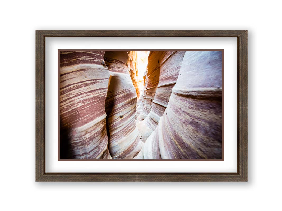 A photo of Zebra Slot Canyon in Escalante National Monument, Utah. A tight formation of alternating, red and white striped stone walls wave into the distance, where yellow light can be seen. Printed on paper, matted, and framed.