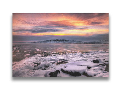 A photo of the sun setting at Mount Timpanogos, Utah, one Christmas evening. Its golden light can be seen peeking through the clouds across the horizon of a frozen lake, wind pushing thin sheets of ice up onto the shore. Printed on canvas.