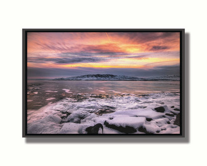 A photo of the sun setting at Mount Timpanogos, Utah, one Christmas evening. Its golden light can be seen peeking through the clouds across the horizon of a frozen lake, wind pushing thin sheets of ice up onto the shore. Printed on canvas in a float frame.