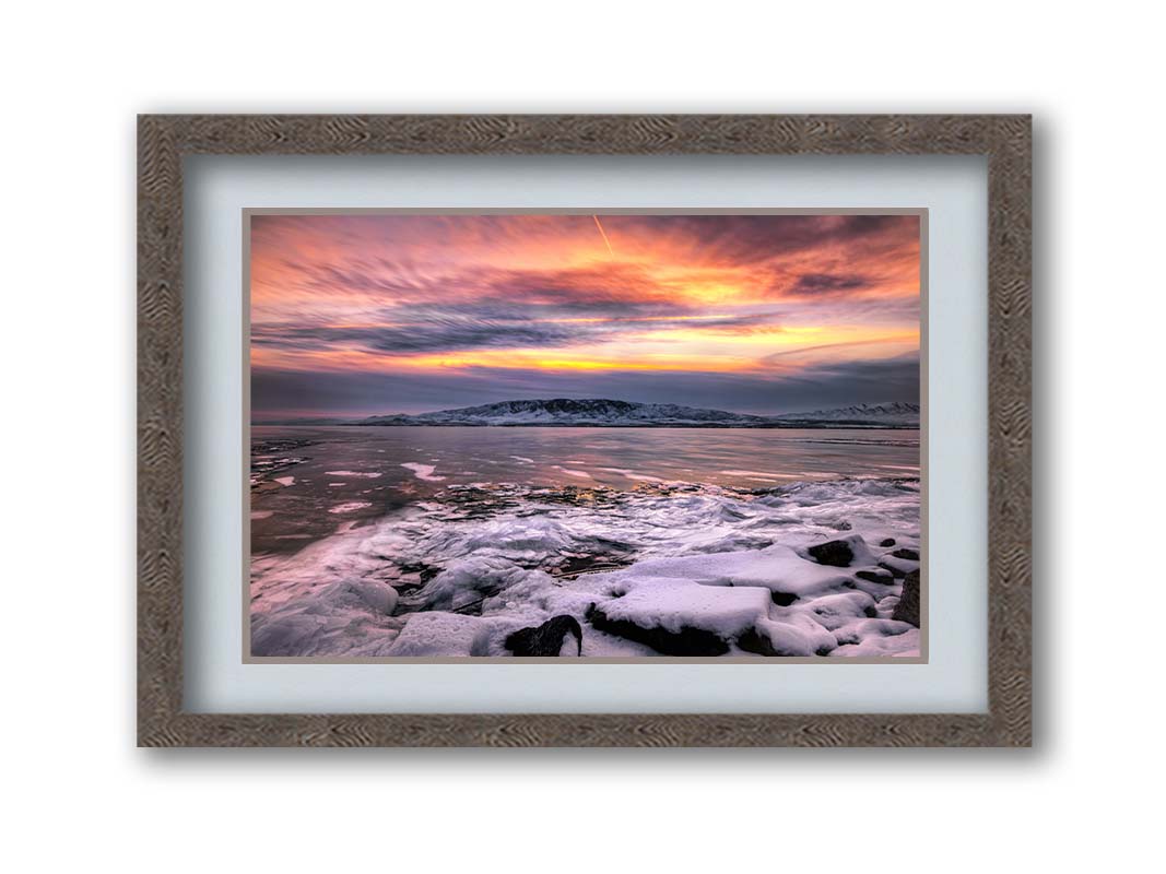 A photo of the sun setting at Mount Timpanogos, Utah, one Christmas evening. Its golden light can be seen peeking through the clouds across the horizon of a frozen lake, wind pushing thin sheets of ice up onto the shore. Printed on paper, matted, and framed.