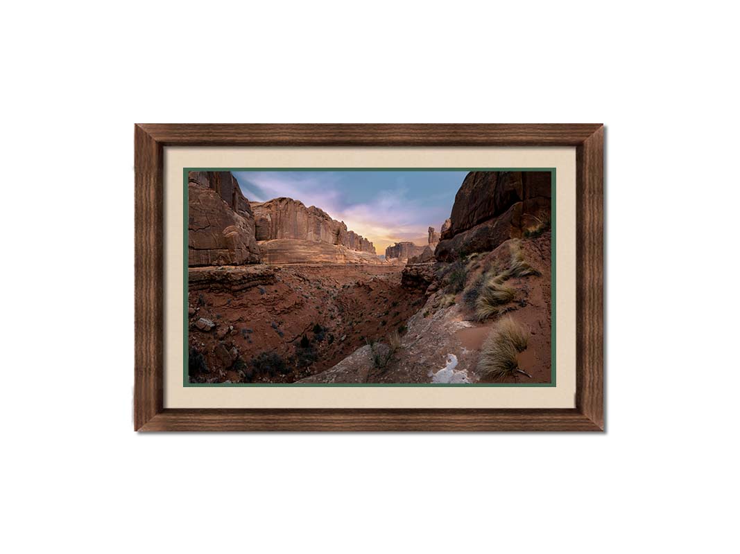 A photo from a Moab, Utah canyon. Sparse shrubbery is scattered along the canyon surrounded by sandstone walls. Printed on paper, matted, and framed.