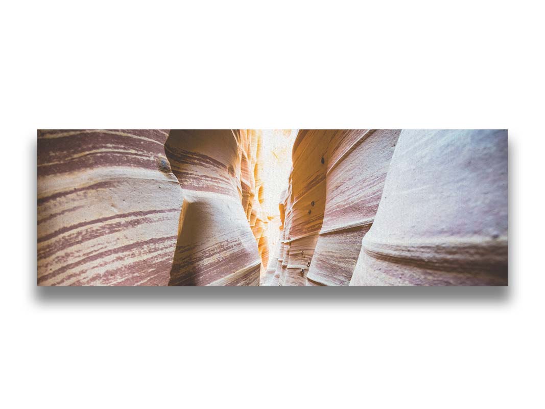 A photo looking down a narrow slot canyon. The walls of sandstone lead out into the light, creating added perspective with their red and white striped layers of stone drawing into the distance. Printed on canvas.