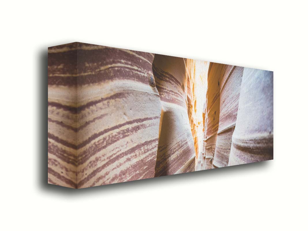 A photo looking down a narrow slot canyon. The walls of sandstone lead out into the light, creating added perspective with their red and white striped layers of stone drawing into the distance. Printed on canvas.