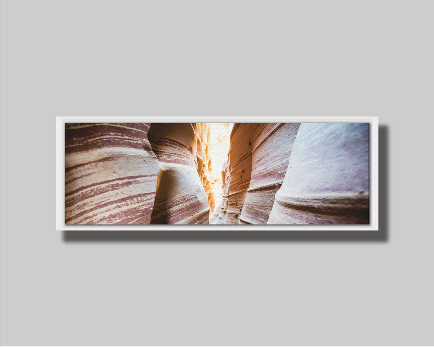 A photo looking down a narrow slot canyon. The walls of sandstone lead out into the light, creating added perspective with their red and white striped layers of stone drawing into the distance. Printed on canvas in a float frame.