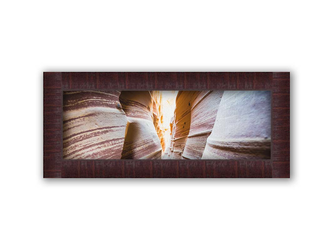 A photo looking down a narrow slot canyon. The walls of sandstone lead out into the light, creating added perspective with their red and white striped layers of stone drawing into the distance. Printed on canvas and framed.