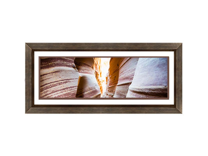 A photo looking down a narrow slot canyon. The walls of sandstone lead out into the light, creating added perspective with their red and white striped layers of stone drawing into the distance. Printed on paper, matted, and framed.