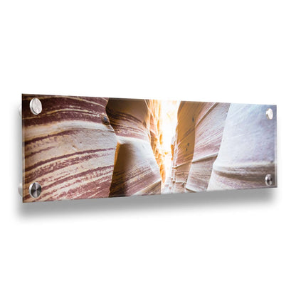 A photo looking down a narrow slot canyon. The walls of sandstone lead out into the light, creating added perspective with their red and white striped layers of stone drawing into the distance. Printed on acrylic.