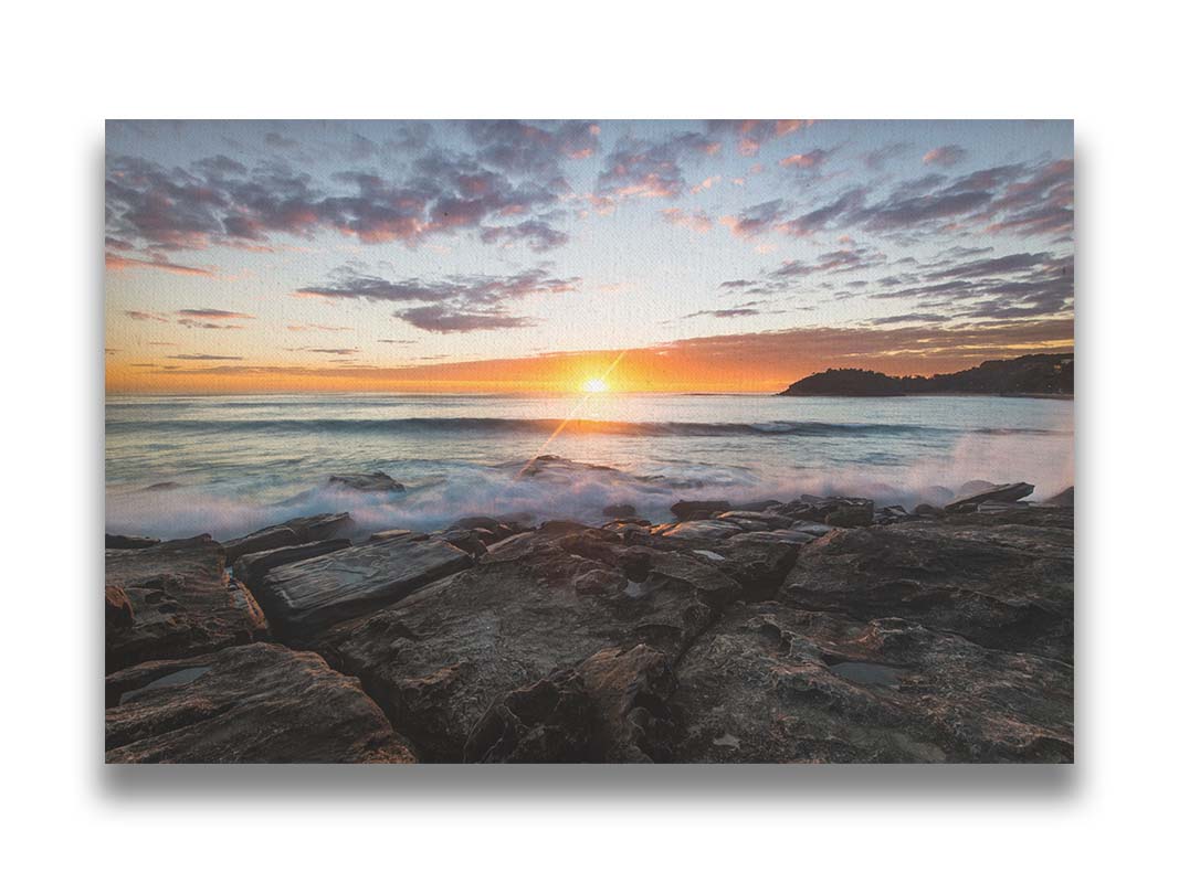 A photo of the sunrise over the ocean horizon at Manly Beach, Sydney, Australia. The sun reflecs brightly off the water as the waves splash against the rocky shore. Printed on canvas.