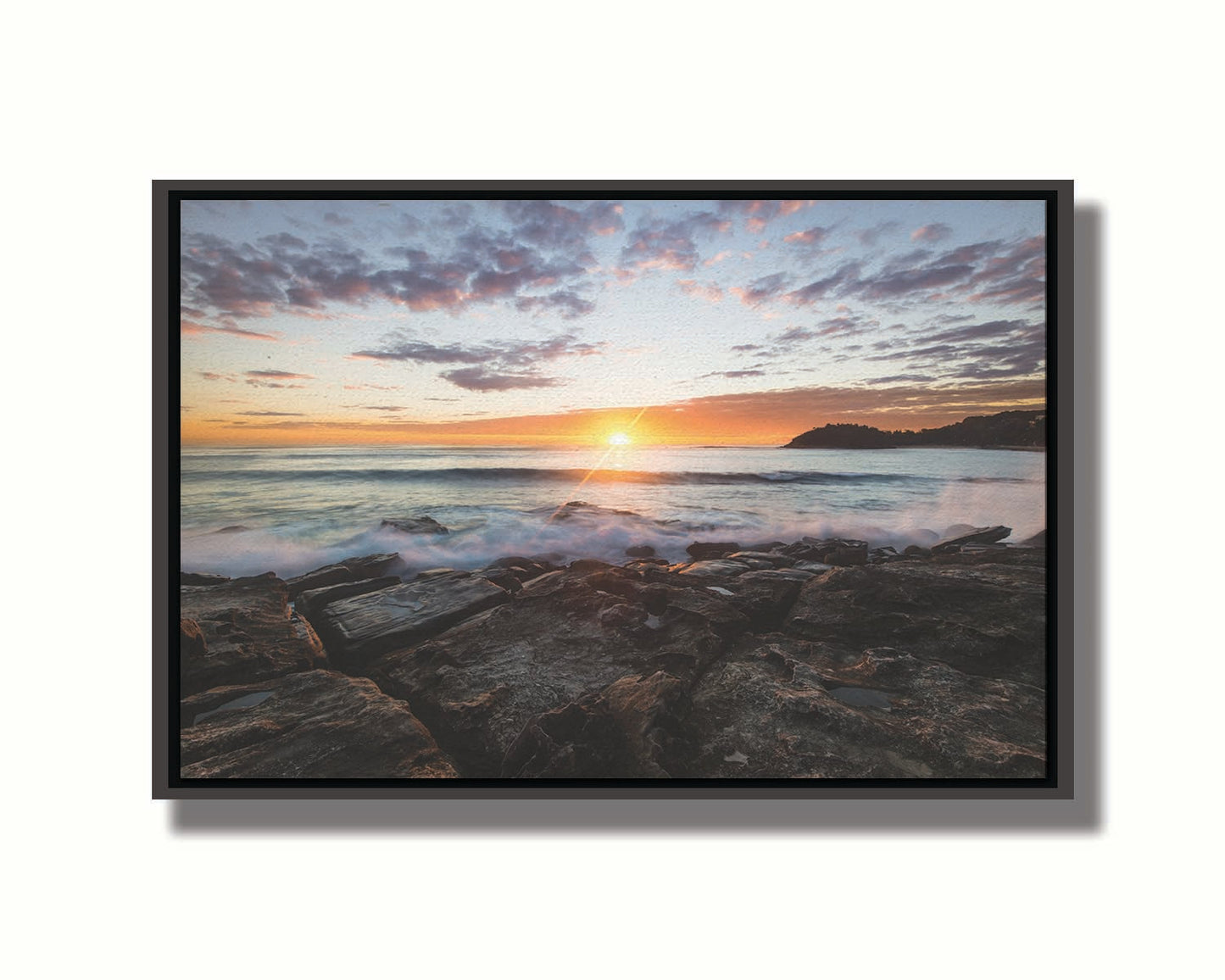 A photo of the sunrise over the ocean horizon at Manly Beach, Sydney, Australia. The sun reflecs brightly off the water as the waves splash against the rocky shore. Printed on canvas in a float frame.