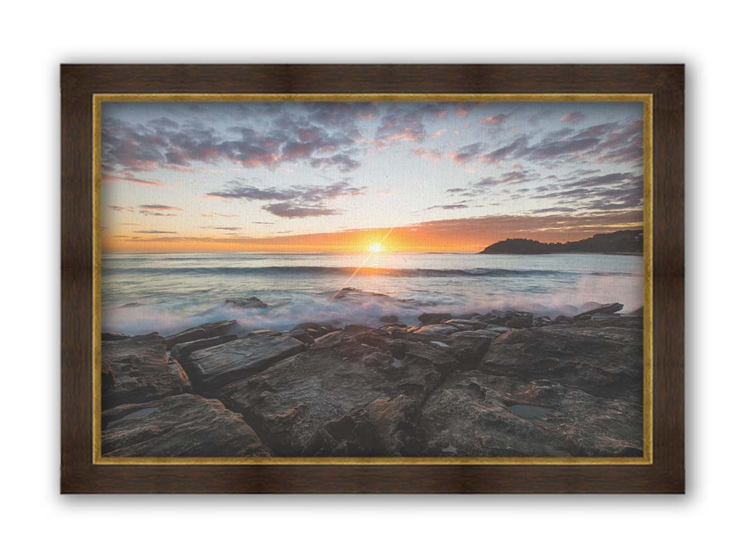 A photo of the sunrise over the ocean horizon at Manly Beach, Sydney, Australia. The sun reflecs brightly off the water as the waves splash against the rocky shore. Printed on canvas and framed.