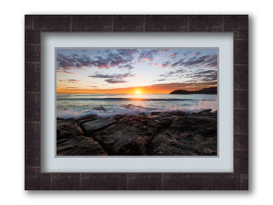 A photo of the sunrise over the ocean horizon at Manly Beach, Sydney, Australia. The sun reflecs brightly off the water as the waves splash against the rocky shore. Printed on paper, matted, and framed.