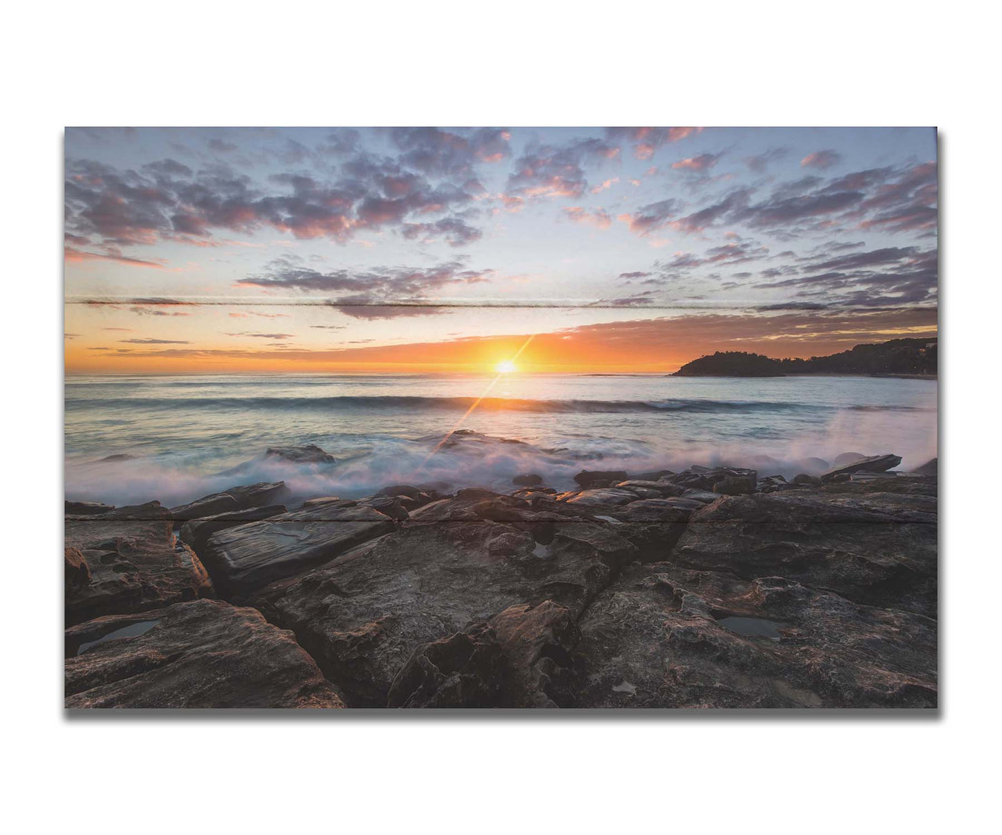 A photo of the sunrise over the ocean horizon at Manly Beach, Sydney, Australia. The sun reflecs brightly off the water as the waves splash against the rocky shore. Printed on a box board.