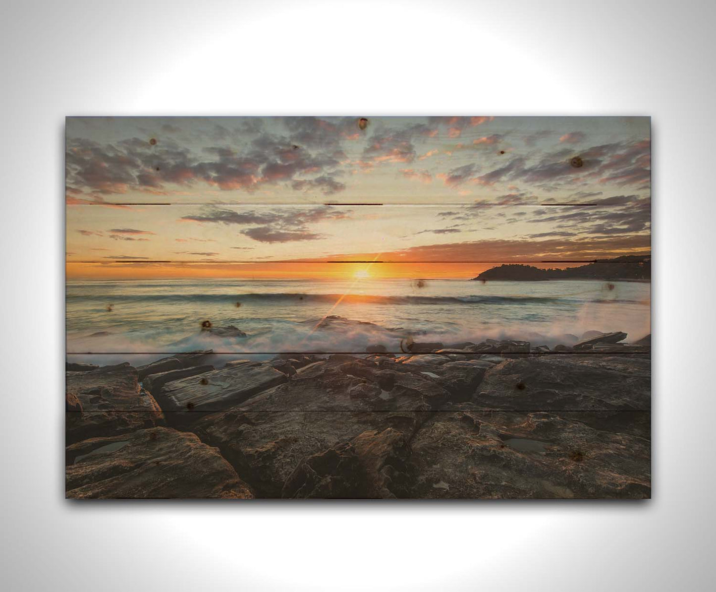 A photo of the sunrise over the ocean horizon at Manly Beach, Sydney, Australia. The sun reflecs brightly off the water as the waves splash against the rocky shore. Printed on a wood pallet.