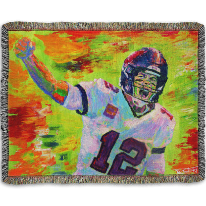 A woven throw pillow designed with a painting of American football player Tom Brady yelling with his fist in the air, rallying his teammates. Arbitrary colors and visible brushstrokes add to the energy of this painting, alongside the bright greens, reds, and oranges of the backdrop.