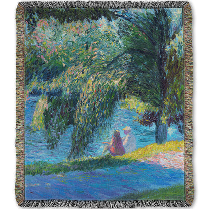 A woven throw blanket designed with a painting of two people sitting by the water, near a park sidewalk, with a large tree above them.