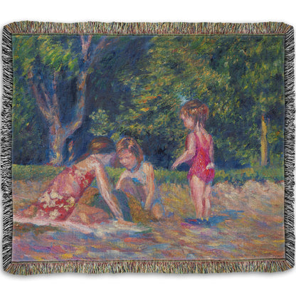 A woven blanket designed with a painting of three children playing in the sand at a beach. Their red swimsuits contrast the green foliage in the background.