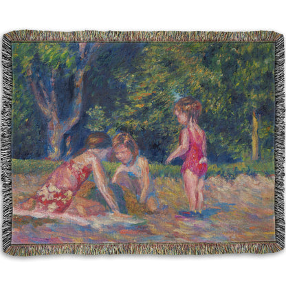 A woven blanket designed with a painting of three children playing in the sand at a beach. Their red swimsuits contrast the green foliage in the background.
