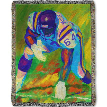 A woven throw blanket designed with a painting of a football player, wearing white and purple, crouched on a bright green field.
