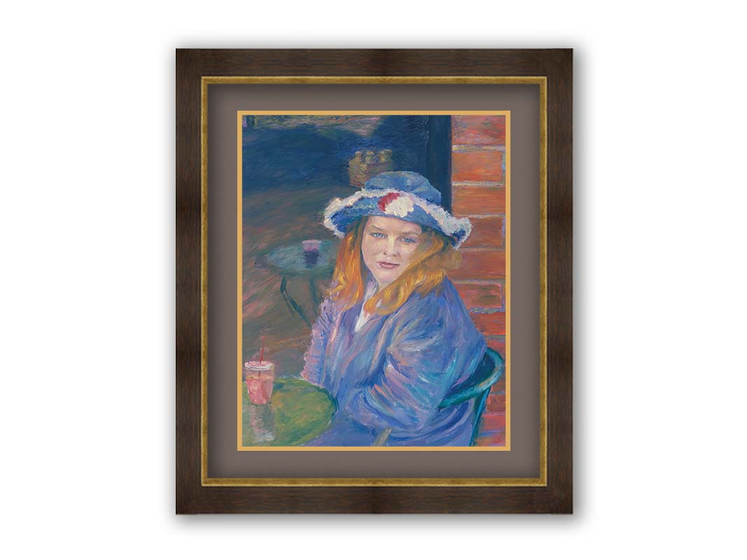 A portrait of a person in a blue outfit, sitting at a table outside a brick building with a drink. Printed on paper, matted, and framed.