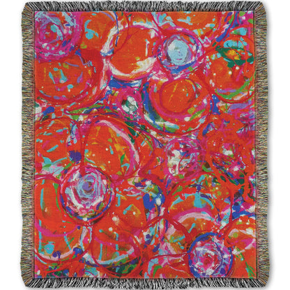 A woven throw blanket designed with a drip painting of a cluster of abstracted flowers created with splatters of orange, red, and pink with accents of green, blue, yellow, and purple.