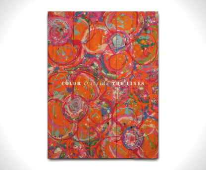 A drip painting of a cluster of abstracted flowers created with splatters of orange, red, and pink with accents of green, blue, yellow, and purple. White text in the center reads "Color outside the lines." Printed on a wood pallet.