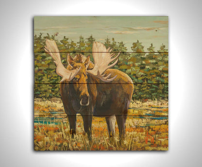 A painting of a moose standing in a bright yellow and orange field, dotted with blue puddles. An evergreen forest borders this Gunflint Trail field in Minnesota. Printed on a wood pallet.