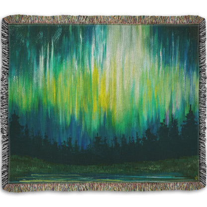 A throw blanket woven into a painting of a colorful aurora in yellow, green, and blue above an evergreen forest.