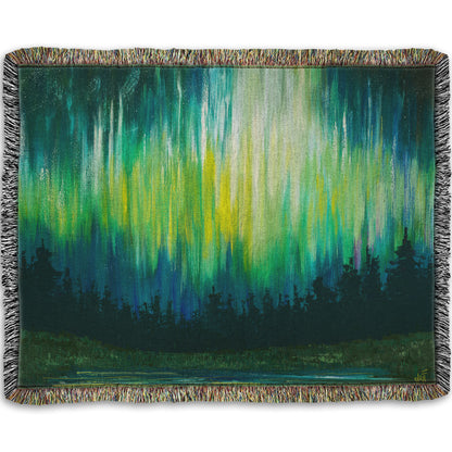 A throw blanket woven into a painting of a colorful aurora in yellow, green, and blue above an evergreen forest.