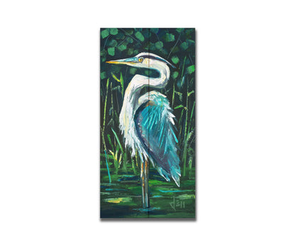 A painting of a great blue heron standing tall in a pond surrounded by vibrant foliage. Printed on a box board.