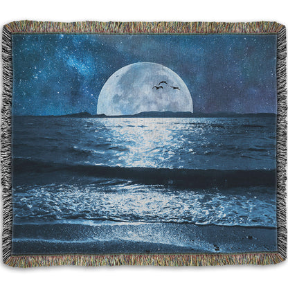 A woven throw blanket designed with a photo of a full moon over the horizon of a calm, dark sea during a starry night.