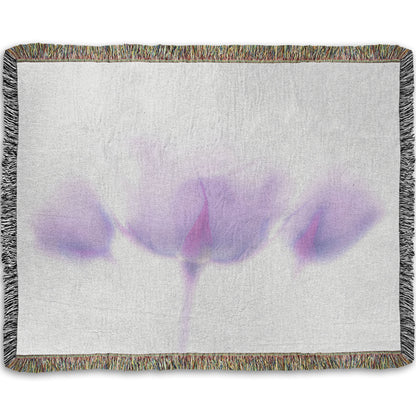 A woven throw blanket designed with a  photo of three pale purple roses against a pure white background, enveloped in a gentle white glow.