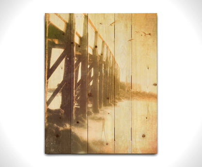 A sepia photo looking out to sea along the side of a pier, as seagulls fly above. Printed on a wood pallet.