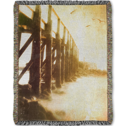 A woven throw blanket designed with a sepia photo looking out to sea along the side of a pier, as seagulls fly above.
