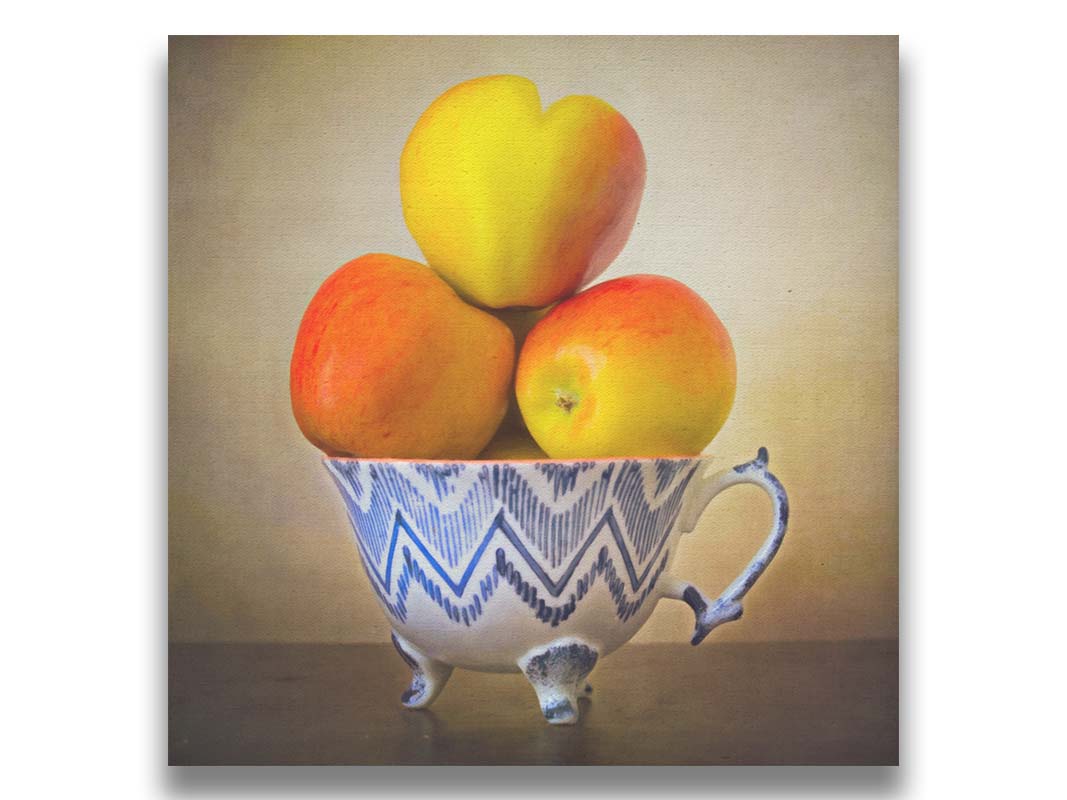 A photograph of a cup full of red and yellow apples, set against a brown table and lighter wall. Printed on canvas.