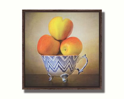 A photograph of a cup full of red and yellow apples, set against a brown table and lighter wall .Printed on canvas in a float frame.