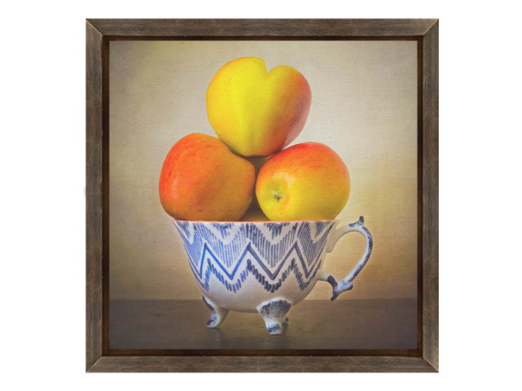 A photograph of a cup full of red and yellow apples, set against a brown table and lighter wall. Printed on canvas and framed.