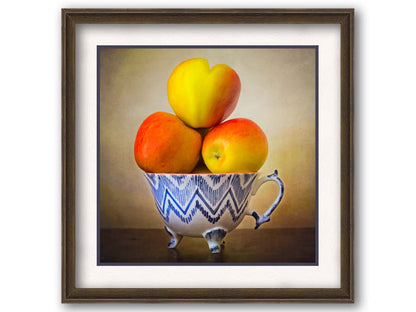  A photograph of a cup full of red and yellow apples, set against a brown table and lighter wall. Printed on paper, matted, and framed.