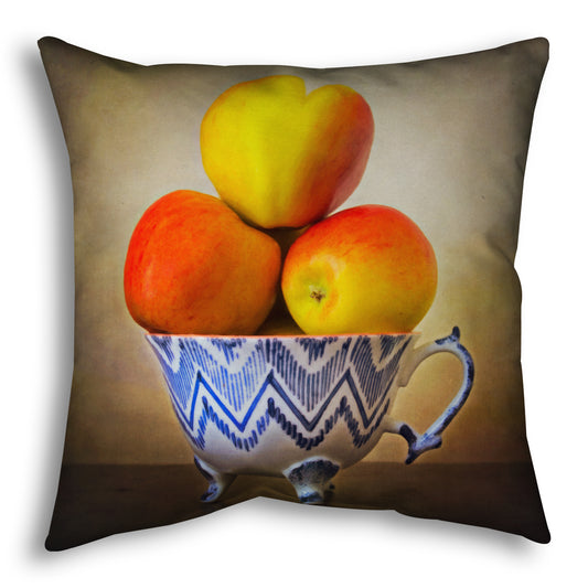 A throw pillow designed with a photograph of a cup full of red and yellow apples, set against a brown table and lighter wall. 