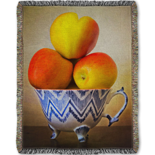 A woven blanket designed with a photograph of a cup full of red and yellow apples, set against a brown table and lighter wall. 
