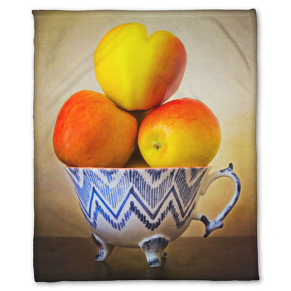 A fleece blanket designed with a photograph of a cup full of red and yellow apples, set against a brown table and lighter wall. 