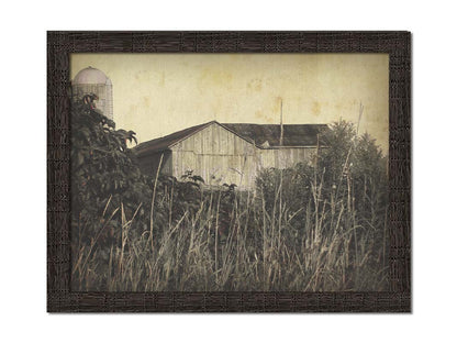 A sepia tone photo of a barn peeking through a foreground of tall grasses and foliage. Printed on canvas and framed.