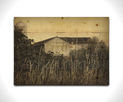 A sepia tone photo of a barn peeking through a foreground of tall grasses and foliage. Printed on a wood pallet.