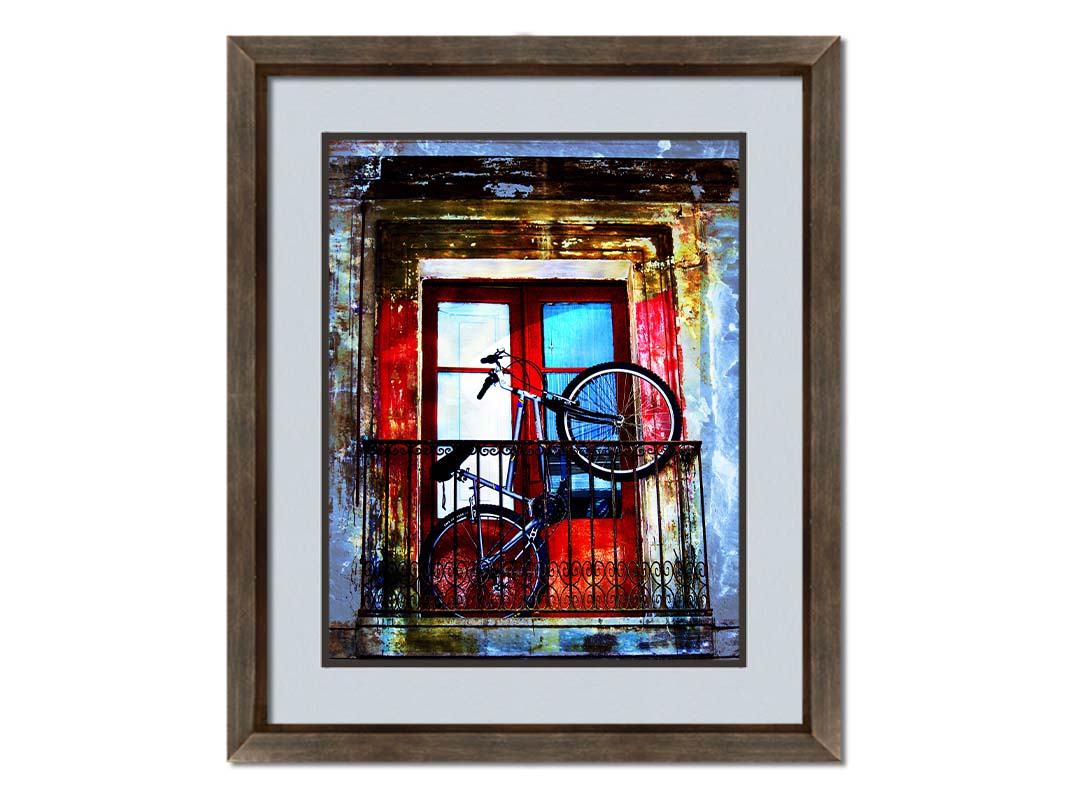 A photo of a bike propped up on a balcony in front of a red door. Printed on paper, matted, and framed.