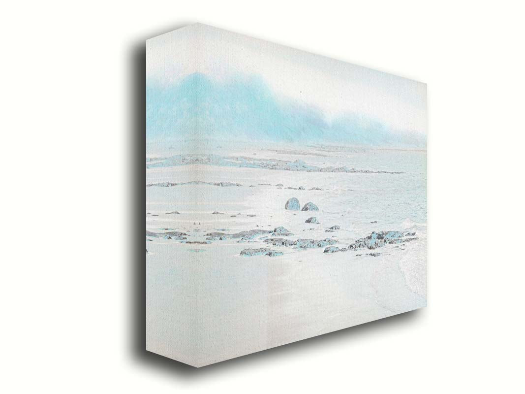 A photo of a rocky beach with a pale blue palette. The sea is calm and washing over the sand and rocks. Printed on canvas.