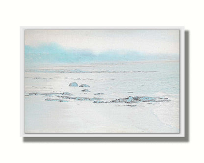 A photo of a rocky beach with a pale blue palette. The sea is calm and washing over the sand and rocks. Printed on canvas in a float frame.