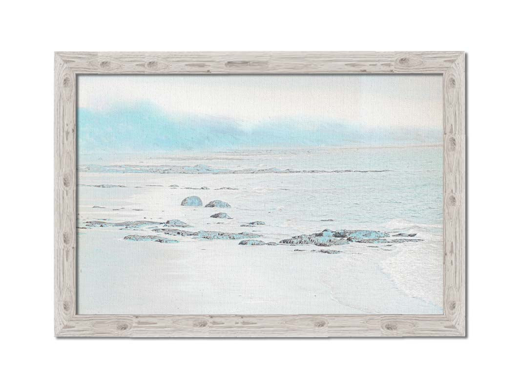 A photo of a rocky beach with a pale blue palette. The sea is calm and washing over the sand and rocks. Printed on canvas and framed.