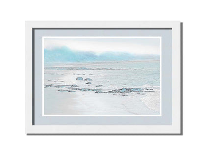 A photo of a rocky beach with a pale blue palette. The sea is calm and washing over the sand and rocks. Printed on paper, matted, and framed.