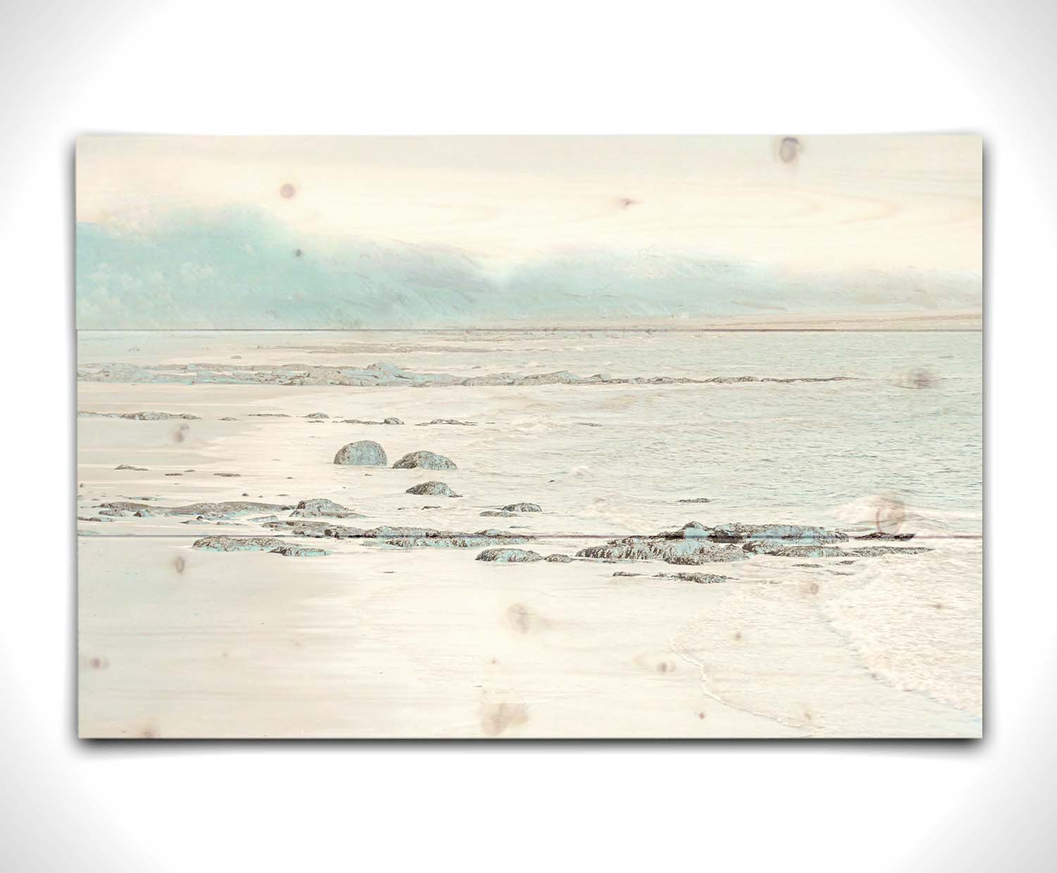 A photo of a rocky beach with a pale blue palette. The sea is calm and washing over the sand and rocks. Printed on a wood pallet.